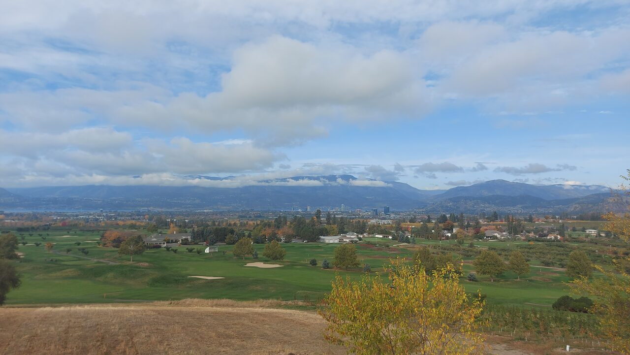 A connected Okanagan working landscape: dry grassland and orchards in the foreground, a city skyline and lake in the valley, and forested mountains beyond.